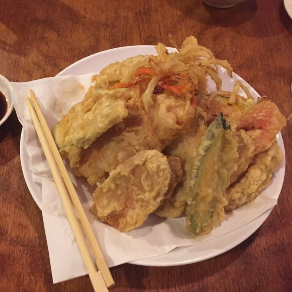 A white plate of golden-brown tempura, including vegetables and shrimp, with chopsticks on a wooden table.